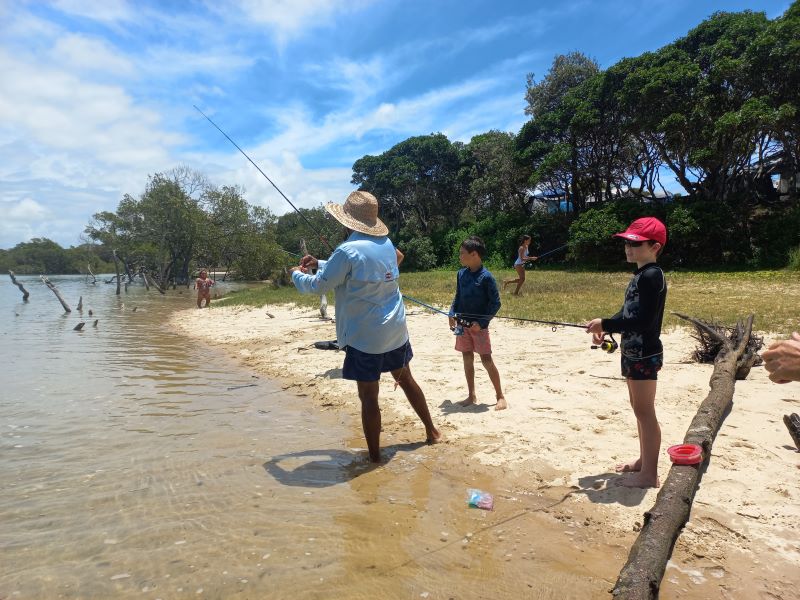 An adult and 2 boys fish on a sandy beach into a river