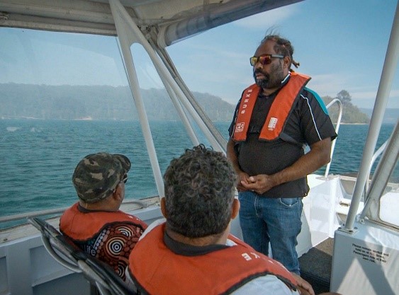 An Indigenous man is standing at the front of a large boat on the ocean, and in front of him are two men seated. He is talking to them as part of his training to become an Indigenous tour guide. 