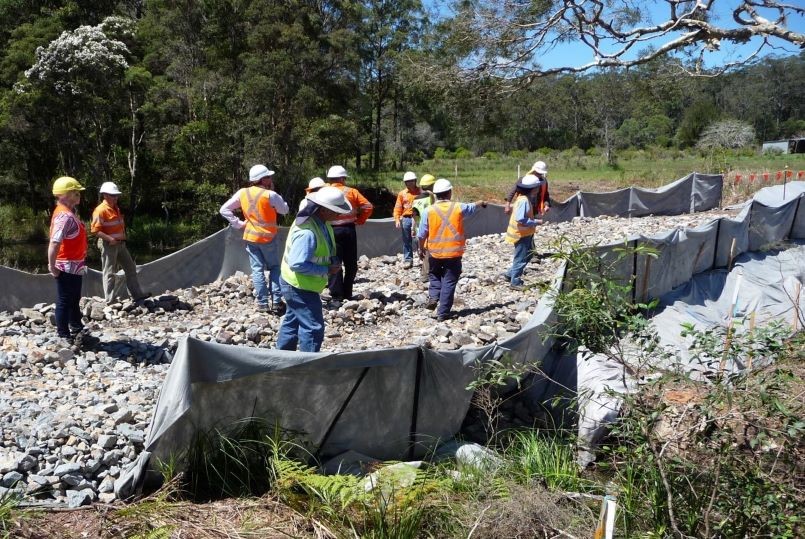 A group of people in hard hats and hi-vis shirts are walking over a construction site in the bush, with fencing to stop sediment run off and lots of blue metal on the ground.
