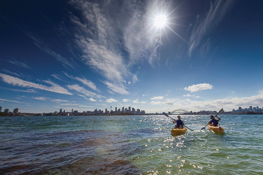 Two people paddling kayaks with the Sydney skyline in the background and a bright sun above.