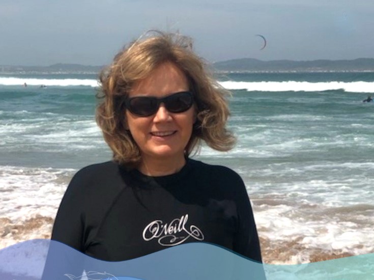A woman in dark sunglasses looks into the camera. The sea is behind her. She is wearing a wetsuit.