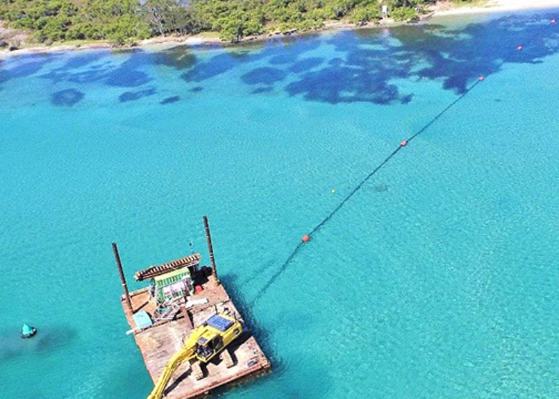 A barge with excavator machine on board floats in clear, blue water next to a tree-lined shore