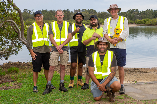 A group photo of six men wearing high visibility vests next to a calm river with a tree-covered riverbank in the background.