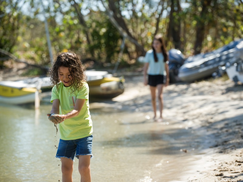 A small boy with long curly hair and a straw hat lifts wet sand from the beach. His sister walks toward him. 