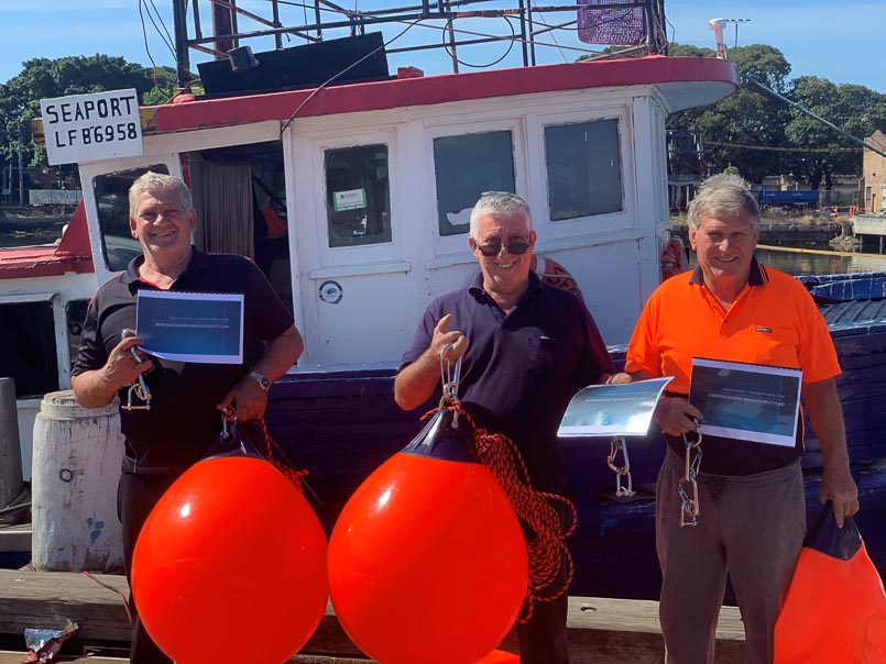 Three men holding booklets and standing next to bright coloured buoys with a fish trawl vessel in the background.