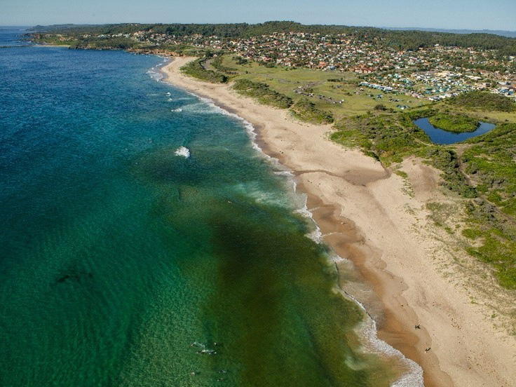 Aerial view of beach with residential and commercial development on the nearby hills. Beautiful bright summer's day with clear water, longboards, surfers and swimmers.