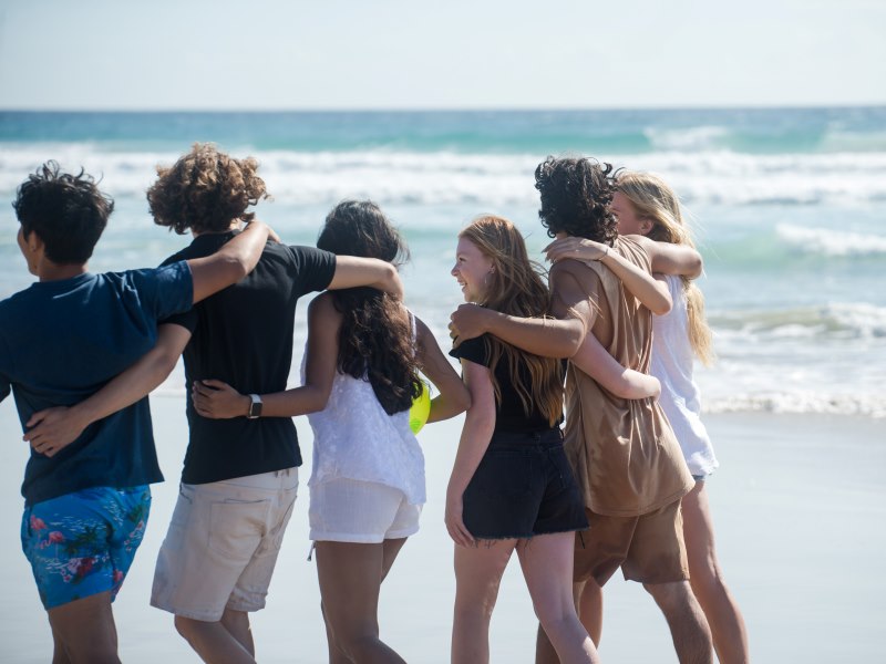 Group of friends walking along the beach arm in arm.  They are smiling and having fun.  One of them is holding a yellow ball.  They are on the edge of the water, walking in the sand. The sky is blue   