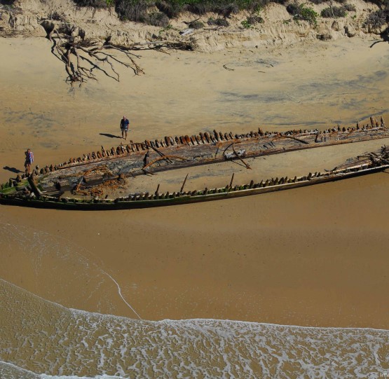 An aerial view of a beach with the outline of boat wreckage visible in the sand between the water and trees.