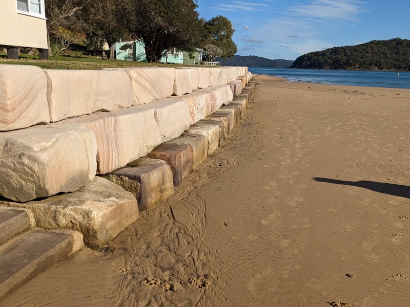 Large sandstone blocks act as a barrier from the land with houses and the river. We see 3 layers of blocks, wet sand leading to a glimpse of water. Above the block breakwater are shack-like houses.