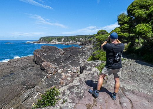 A man is standing on a rock platform while looking through binoculars at ocean and coastline.
