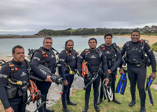 Six men in wetsuits and scuba gear standing on headland in front of a beach