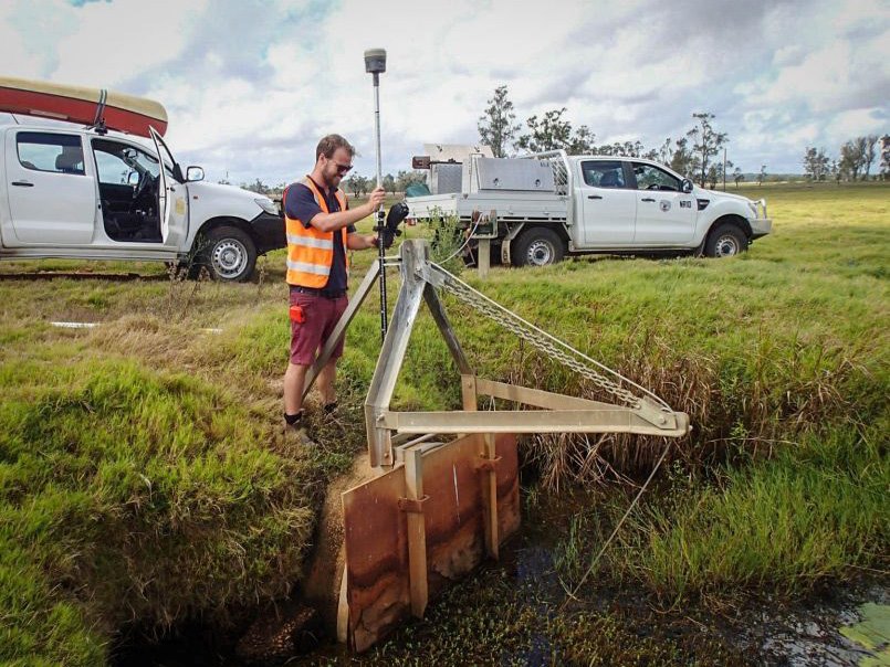 A surveyor in high vis vest holds a surveying tool. He is measuring a metal frame that opens flood gate. We can see a metal gate with chain and pulley that will open the gate. It is closed. In the background are two utes with toolkits. One has a canoe on top. The worker has a beard and sunglasses. He is standing on thick grassed area. The water from the flow looks to be full of weeds and is brackish.