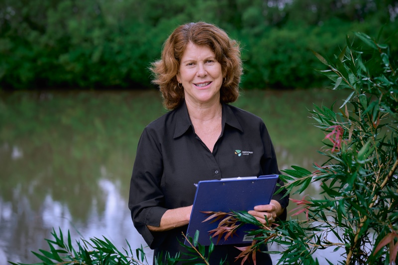 Portrait of female council worker on riverbank with clipboard, smiling at camera. Bushy foliage framing the image, river behind the woman. 