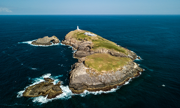 South Solitary Island is a rocky island with a smooth green top marked by a white lighthouse at the furthest peak. All set in a dark blue, calm ocean. 