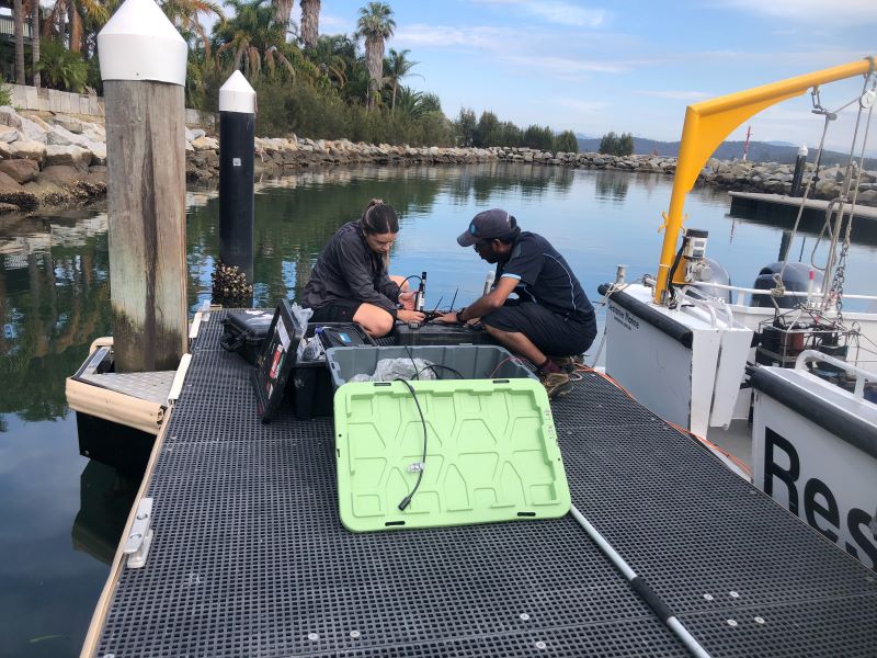 Two scientists kneeling on a jetty in a New South Wales marina, next to a moored boat, with open cases full of electronic equipment on their shoreward side and towards the camera. They are intently focused on a spectral analyser that sits between them. The water behind them is very calm and reflects the palm trees that line the shore. A seawall of sandstone blocks encircling the marina is visible in the background.