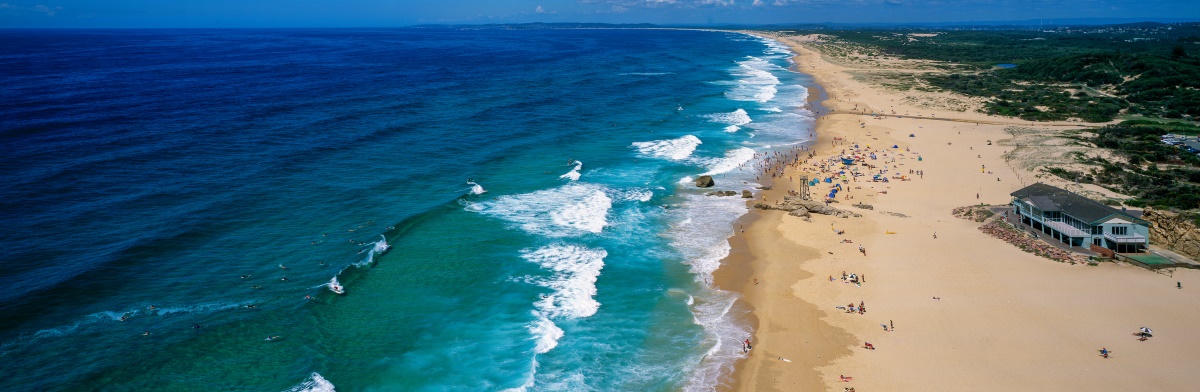 Distant view of beach during a bright summer’s day with lots of sunbathers, swimmers and surfers. Buildings and concrete boardwalks adjacent to the beach sand.