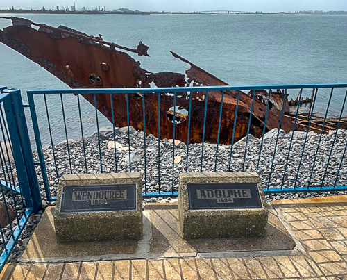 Two plaques sit at ground level in front of a metal railing and overlooking the rusted remains of a boat with water and a distant city skyline in the background.