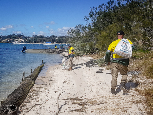 Two mean wearing high visibility shirts are on a sandy beach and placing rubbish in large white bags. A boat is at anchor in the background.