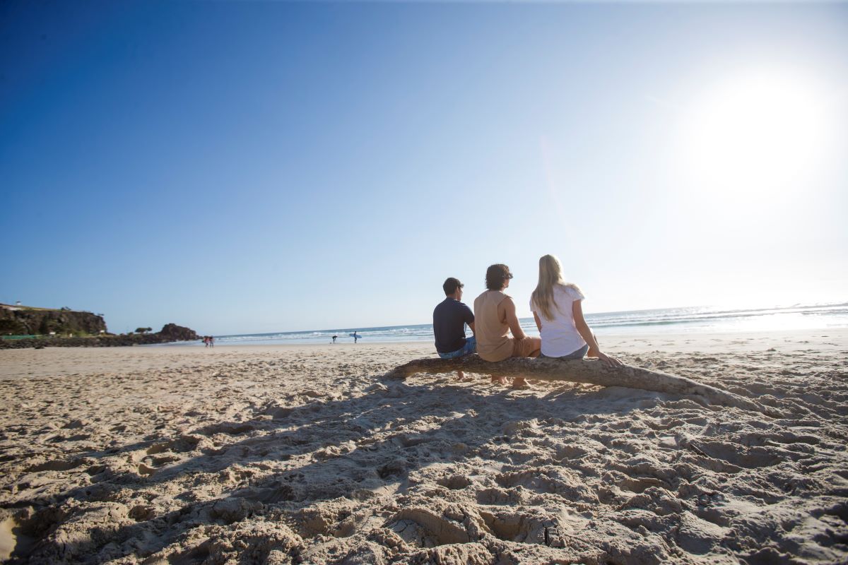  Alt text: 4 teenagers sit on a log facing the ocean, they are watching the sun rise. 