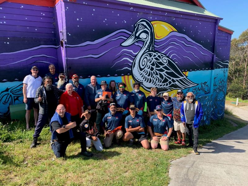 Group of people pose in front of a building that has been painted with a colourful artwork depicting a native duck, mountains, stars and water. The art of showing water shows marine creatures.